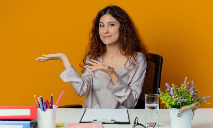 pleased-young-pretty-female-office-worker-sitting-desk-with-office-tools-points-with-hands-side-isolated-orange_141793-73519